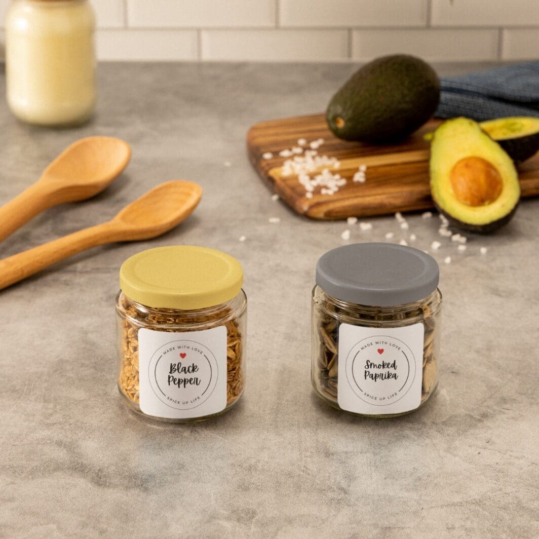 This image features a well-organized kitchen setup showcasing jars filled with spices. Jars are labeled "Black Pepper" and other 75 names of spices, peppers, and herbs, and can be places on your kitchen countertop. They are available uniquely at SharekknaOnline.com. The jars labels can be downloaded as png with transparent background, or as pdf with 8 labels per page, and can be printed at home or your local print house.