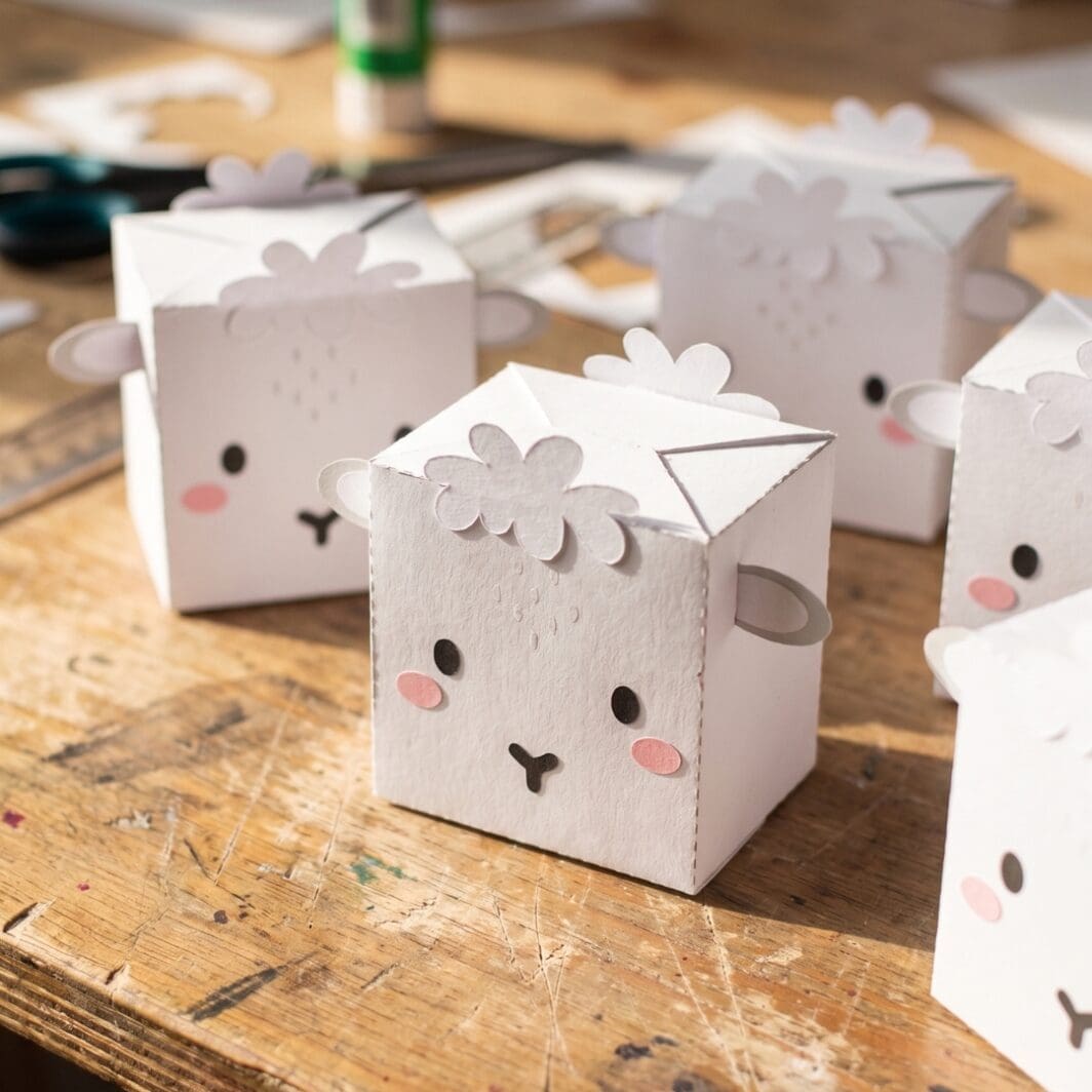 Printable “Let’s Build a Sheep” DIY craft cover image showing a cute assembled sheep paper box with a smiling face, pink cheeks, and attached ears and wool pieces, alongside the flat template with cut and fold lines for easy assembly.